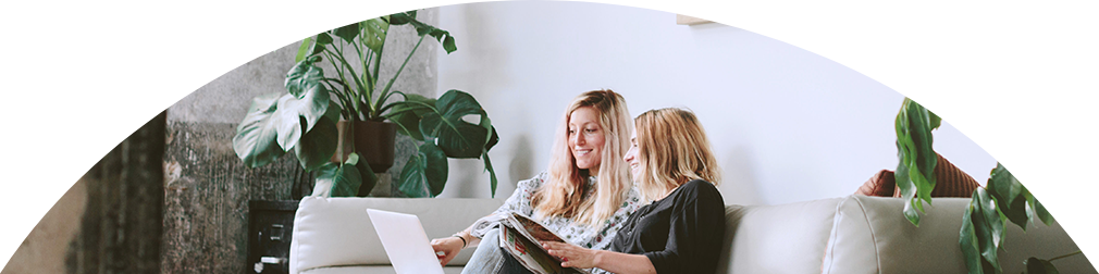 Zwei Frauen sitzen am Sofa und betrachten etwas am Laptop.