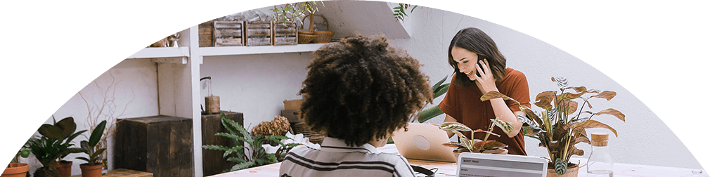 Zwei Frauen sitzen an einem Schreibtisch und arbeiten am Laptop. Eine telefoniert.