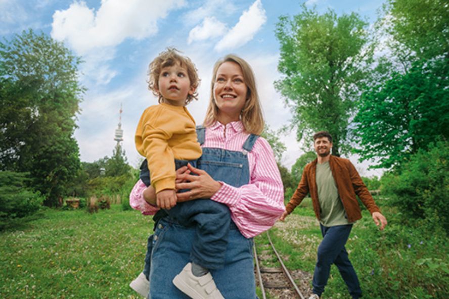 Eine junge Mutter steht mit einem Kleinkind am Arm auf einer Wiese - im Hintergrund kommt der Vater dazu